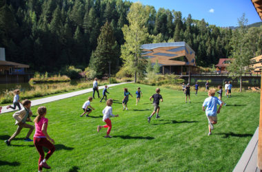 A group of children playing in a lush green field surrounded by pine trees and blue skies at Aspen Country Day School, an independent school in Aspen, Colorado.